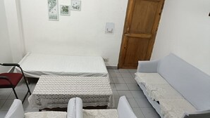 Een kleine, netjes ingerichte kamer met een bed, een slaapbank en een stoel. Er is een houten deur en een stopcontact.