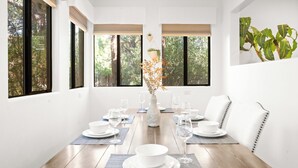 Une salle à manger avec une table en bois dressée pour six personnes, des chaises blanches et un vase contenant des fleurs séchées.