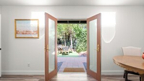 Une porte ouverte donnant sur un patio avec une table et des chaises, un tableau encadré au mur et un plancher en bois à l’intérieur.