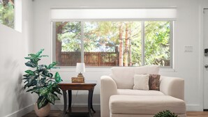 Un salon avec un canapé beige, une table basse en bois, une plante en pot et une vue sur une clôture en bois et des arbres, visible à travers une grande fenêtre.