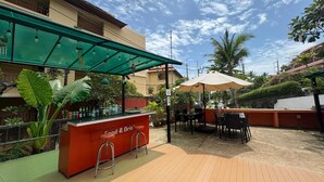 Un espace bar extérieur doté d’un comptoir portant l’inscription « Nourriture et boissons », entouré de tables et de chaises, et abrité par une tonnelle verdoyante.