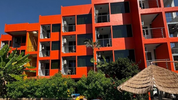 Une piscine à l’eau d’un bleu cristallin, entourée d’une terrasse en bois. Au fond, on aperçoit un immeuble à plusieurs étages, doté de balcons et d’une façade rouge.