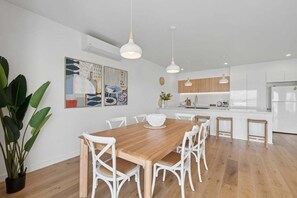 A modern kitchen with white cabinets, a wooden dining table, and hanging pendant lights. - Sandy Toes - Walk to Beach, Pet Friendly, Stylish (Woolgoolga)