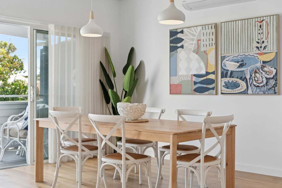 Un coin repas avec une table en bois et des chaises blanches, un vase blanc et des œuvres d’art abstraites accrochées au mur.