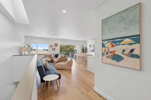A spacious living area with a long bench, a sofa, and a dining table. There is a painting of a beach scene on the wall. - Sandy Toes - Walk to Beach, Pet Friendly, Stylish (Woolgoolga)