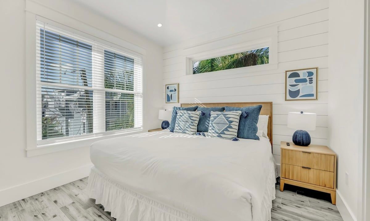 A bedroom with a large bed, a nightstand, a window with blinds, and wall art.