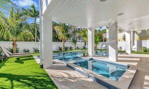 Ein moderner Poolbereich im Freien mit überdachter Terrasse, Palmen und einem gepflegten Rasen.