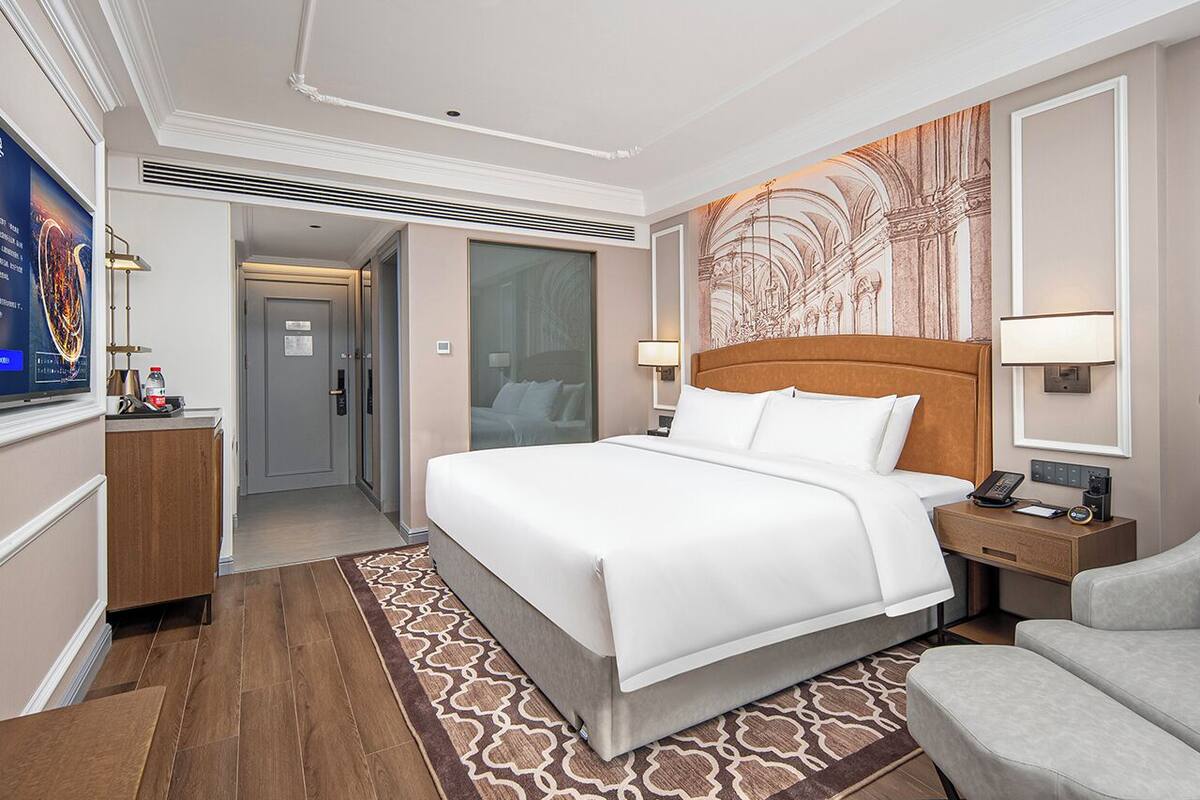 Une chambre d’hôtel avec un grand lit, une table de chevet en bois, un bureau et une chaise. On y trouve une porte donnant sur une autre pièce et un mur orné d’une fresque décorative.