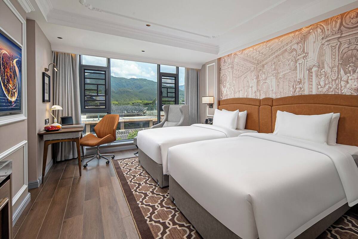 Une chambre d’hôtel avec deux lits, un bureau et une chaise. Une grande fenêtre offre une vue sur les montagnes.