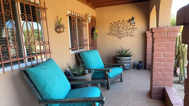 A patio with two wooden chairs with teal cushions, a decorative wall piece, and a cactus in the background. - Enjoyable 2-bedroom house with AC in Green Valley, Nice covered patio. (Green Valley)