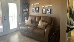 A brown leather sofa with two cushions, a bookshelf with decorative items, and two framed pictures on the wall. - Enjoyable 2-bedroom house with AC in Green Valley, Nice covered patio. (Green Valley)