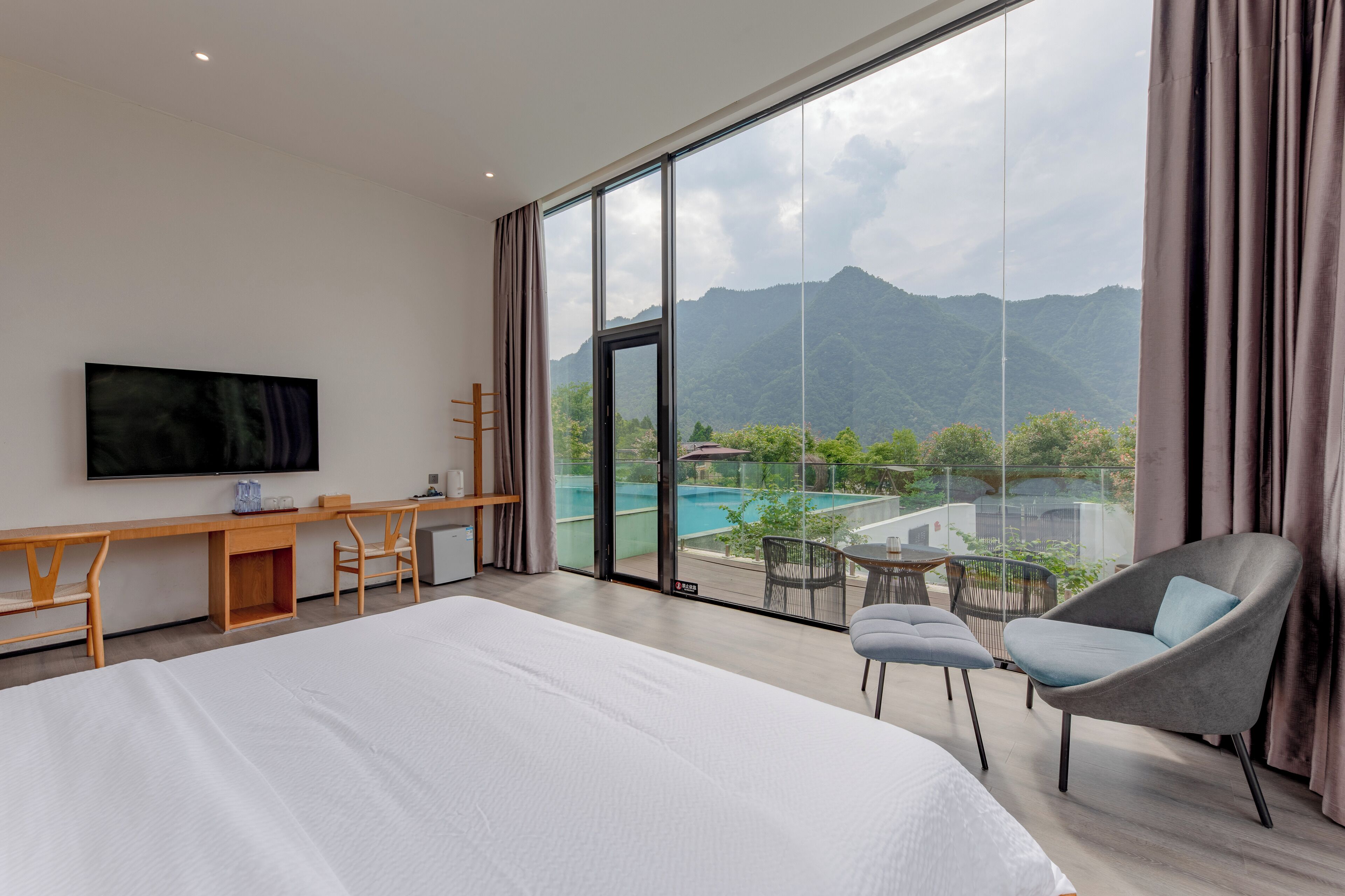 Habitación de hotel con una cama grande, un escritorio, una silla, televisión y balcón con piscina y vistas a la montaña.
