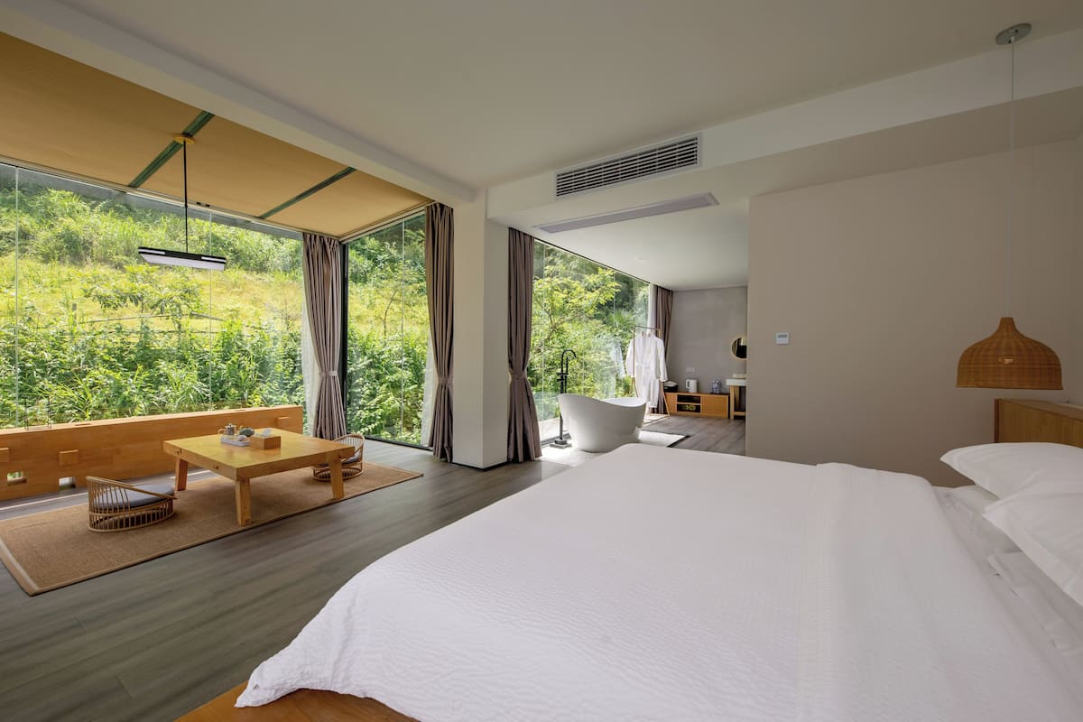 Ein modernes Hotelzimmer mit einem großen Bett, einem hölzernen Couchtisch, einer Sitzecke und Blick auf Grün hinter großen Fenstern.