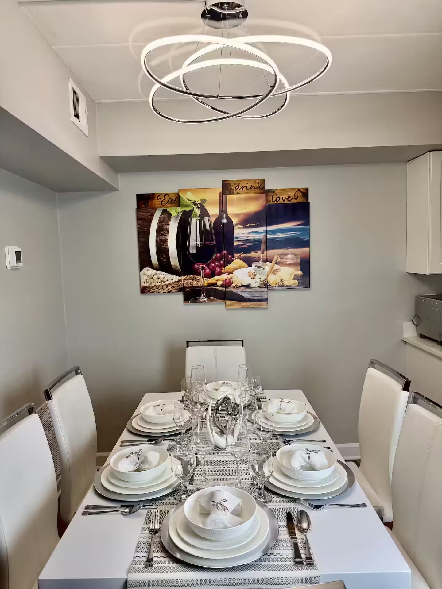 Een eetruimte met een witte tafel voor zes personen, een moderne kroonluchter en een wand met drie ingekaderde afbeeldingen van eten en drinken.