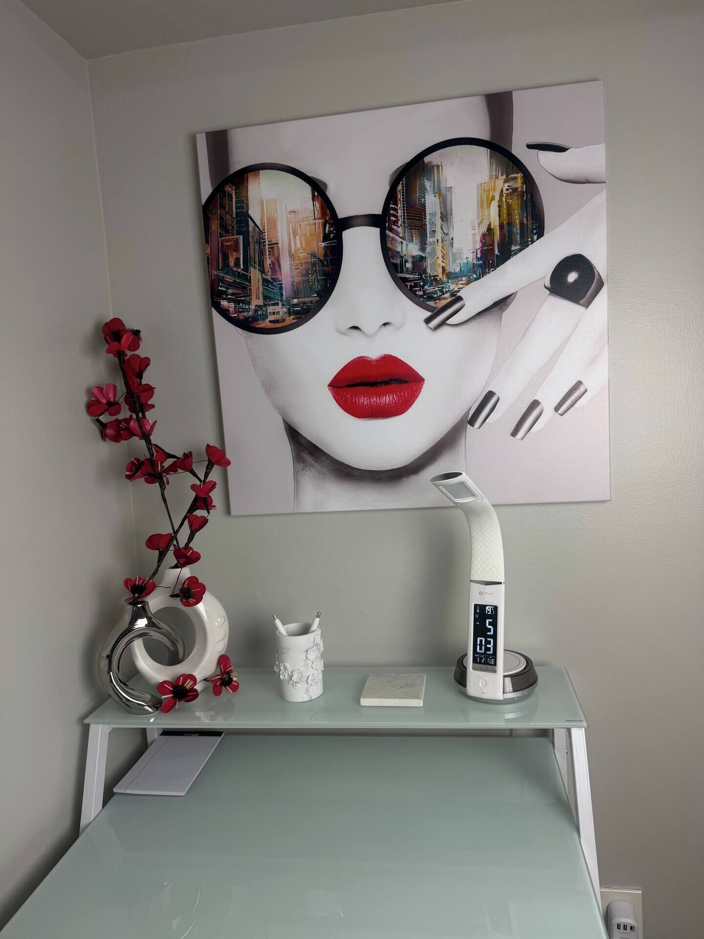 Een moderne badkamer met een glazen bureau, een digitale klok, een vaas met rode bloemen en een kunstwerk in een lijst aan de muur.