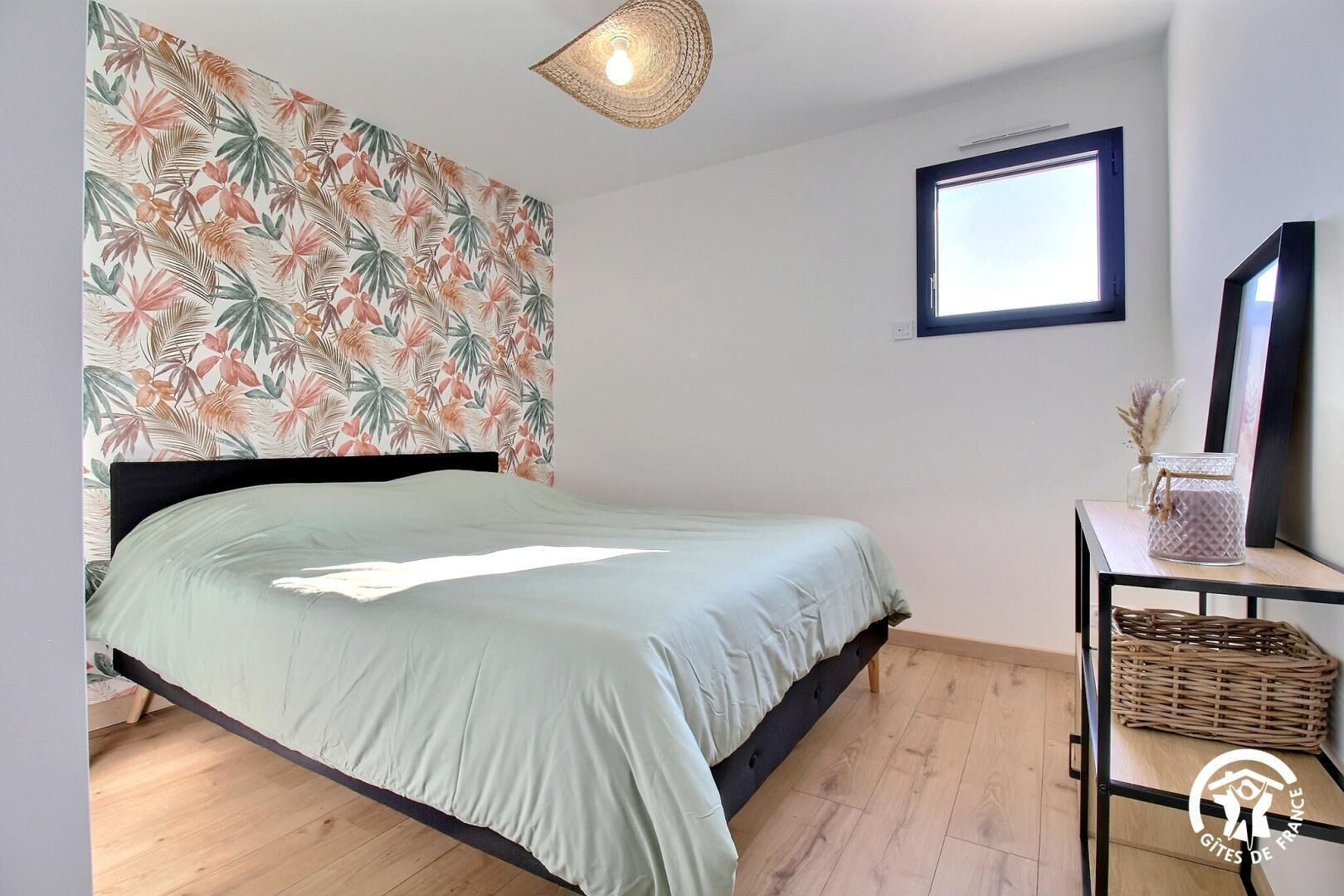 Une chambre à coucher avec un lit, une fenêtre, un miroir fixé au mur et un panier en osier posé sur une table.