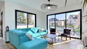Ein modernes Wohnzimmer mit einem türkisfarbenen Sofa, einem Glastisch und einem schwarzen Sessel. Große Fenster bieten einen Ausblick nach draußen.