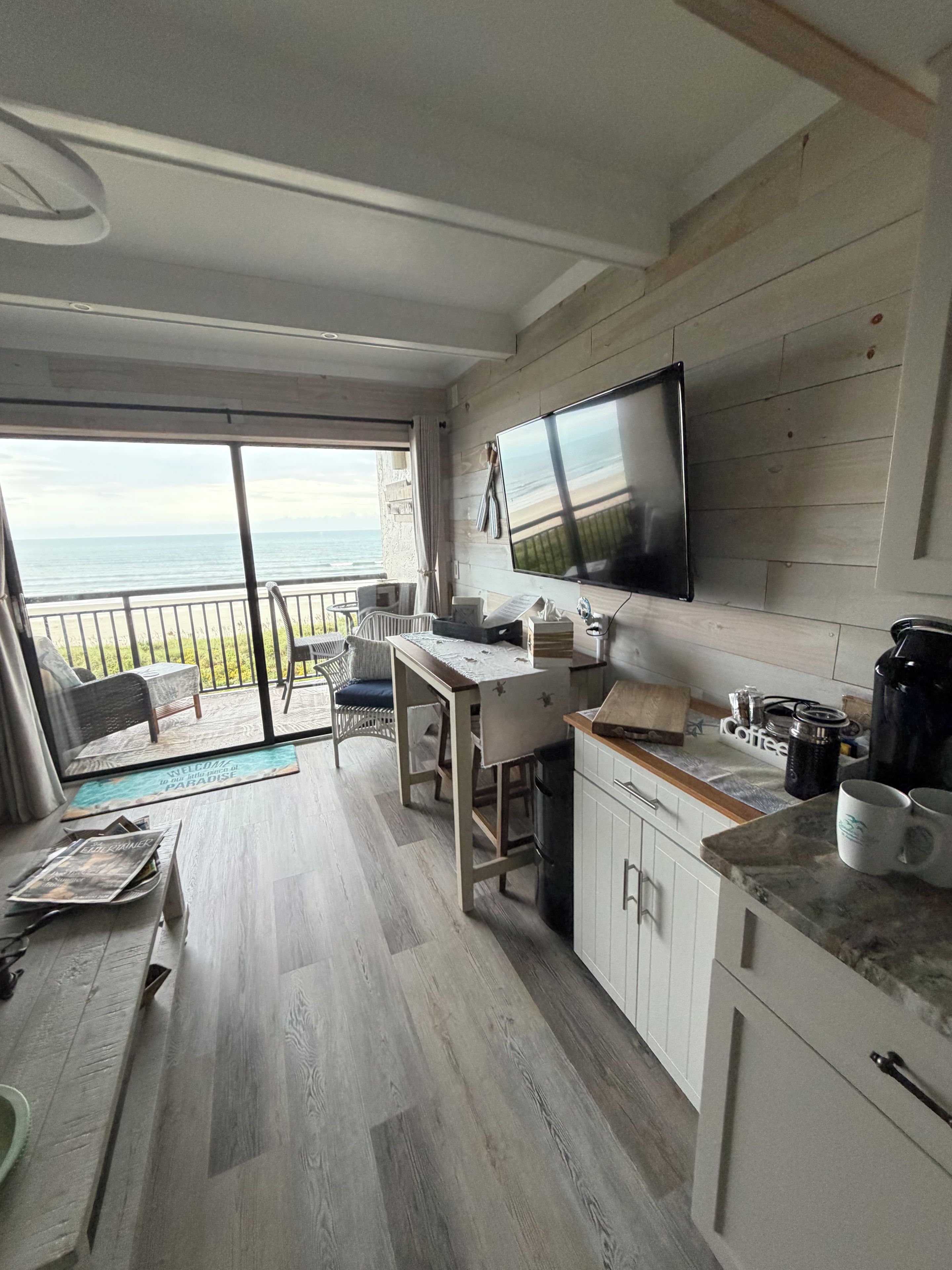 A modern kitchen with a large flat-screen TV, wooden flooring, and a view of the sea through a sliding glass door.