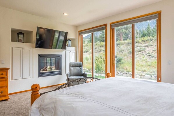 Quarto com cama grande, lareira, TV fixada acima e vista para paisagem montanhosa através de grandes portas de vidro deslizantes.