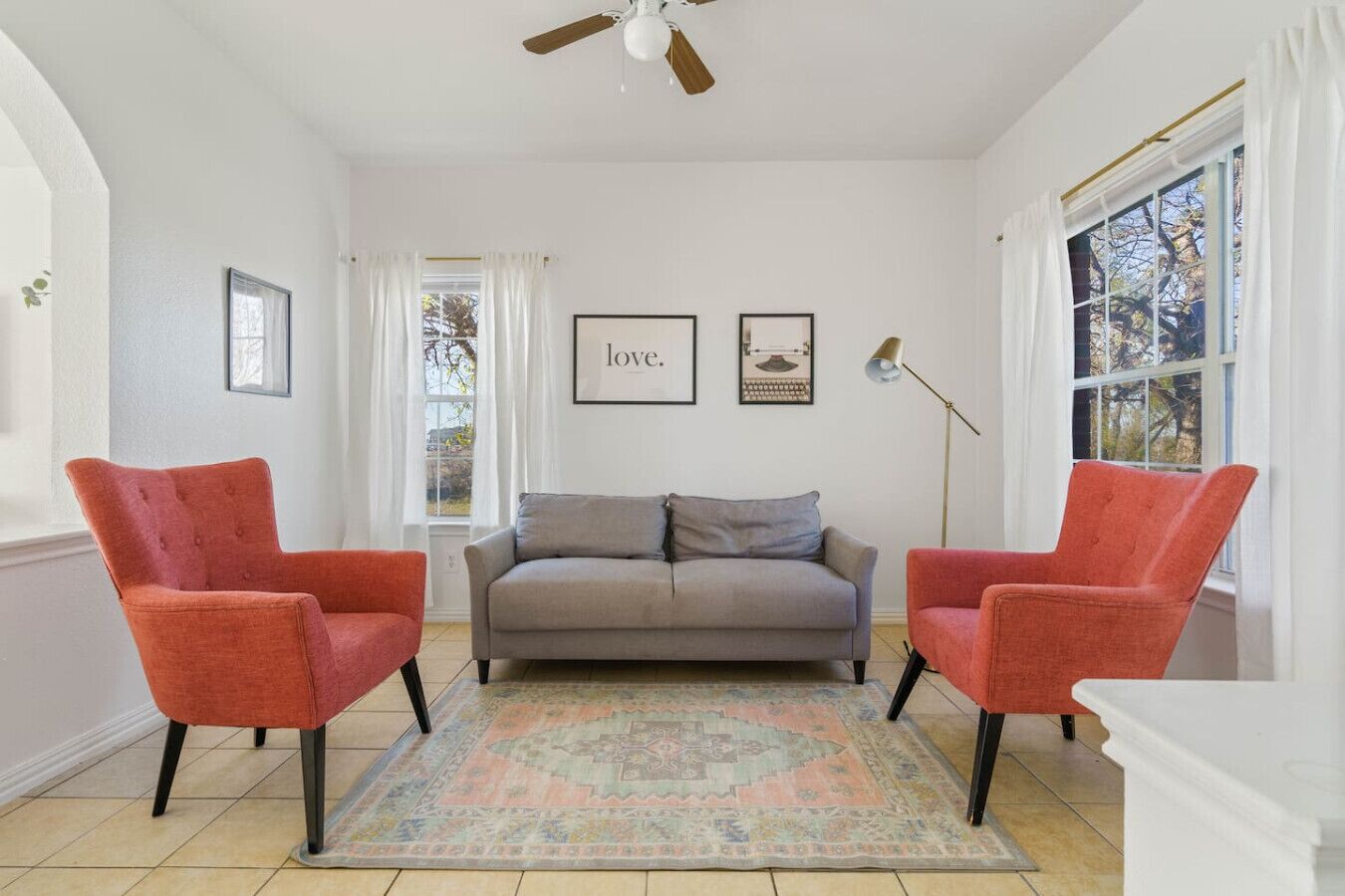 A living room with a grey sofa, two red armchairs, a patterned rug, a ceiling fan, and framed artwork on the wall.
