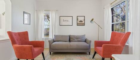 A living room with a grey sofa, two red armchairs, a patterned rug, a ceiling fan, and framed artwork on the wall.