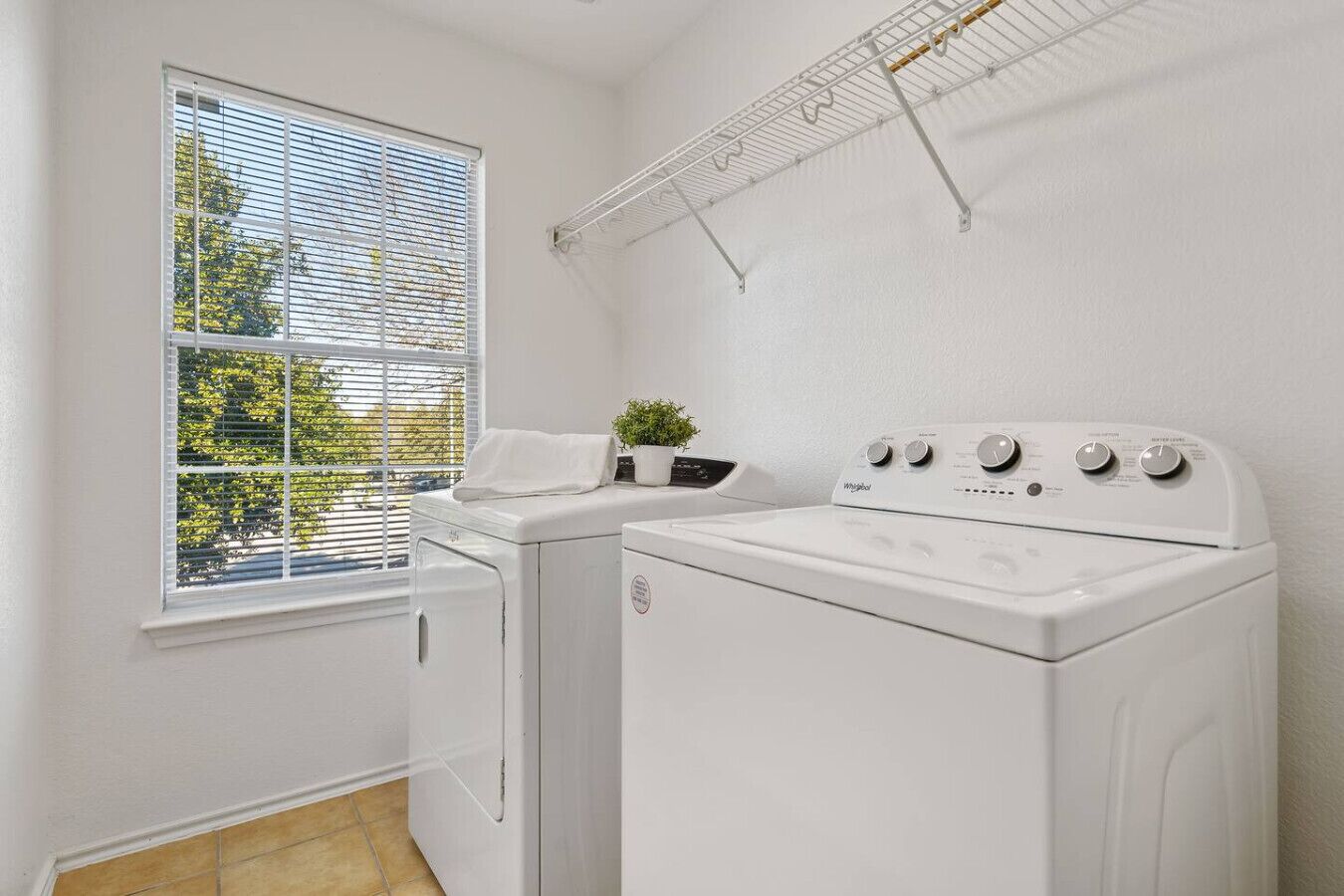 A laundry room with two white front-loading washers and dryers, a drying rack, and a window with blinds.