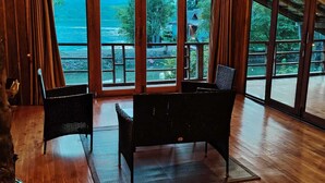 A room with a large sliding glass door leading to a balcony with a view of a body of water and greenery. There are two wicker chairs and a small table in the room. - Nirvana Archipel Resort (Vang Vieng)