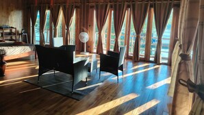 A wooden room with a bed, two chairs, a standing fan, and large windows with curtains. - Nirvana Archipel Resort (Vang Vieng)