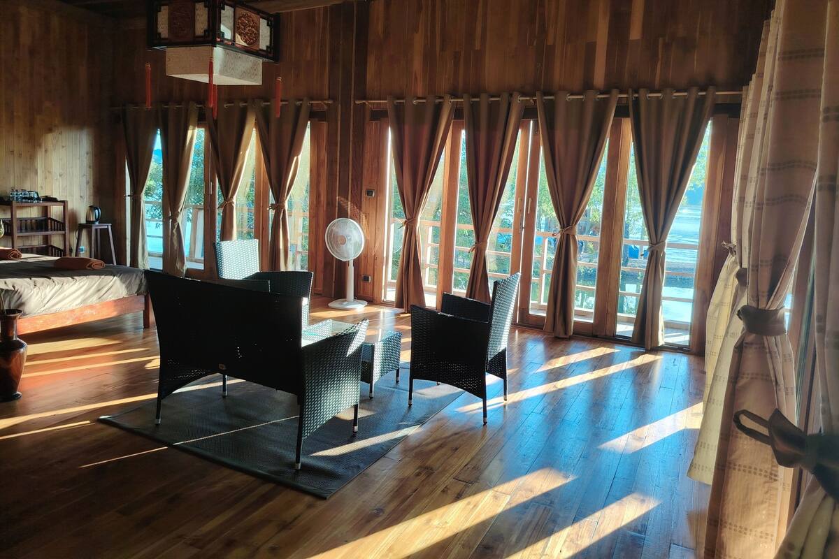 A wooden room with a bed, two chairs, a standing fan, and large windows with curtains.