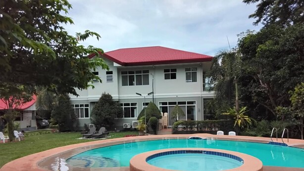 Una piscina con un jacuzzi circular, rodeado de un jardín ajardinado y un edificio de techo rojo al fondo.