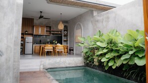 Una cocina moderna con armarios de madera, un comedor con sillas, un ventilador de techo y una pequeña zona de piscina con plantas.