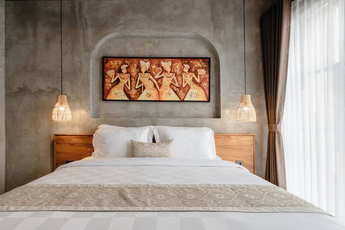 Una habitación de hotel moderna con una cama grande, un cabecero de madera, dos lámparas fijadas a la pared y una obra de arte grande enmarcadas sobre la cama.