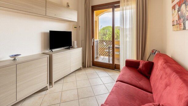 A room with a TV on a wooden cabinet, a red sofa, and a sliding glass door leading to a balcony. - Apartment With air Conditioning and sea Views (Castelsardo)