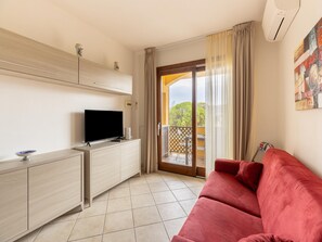 A room with a TV on a wooden cabinet, a red sofa, and a sliding glass door leading to a balcony. - Apartment With air Conditioning and sea Views (Castelsardo)