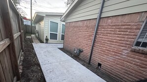 Ein schmaler Betonweg führt zu einem Haus mit Backsteinfassade und einem Holzzaun an einer Seite.