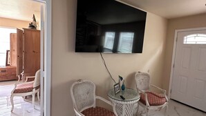 Ein Wohnzimmer mit einem an der Wand montierten Flachbildfernseher, umgeben von drei Korbstühlen mit gemusterten Kissen. Der Raum hat einen gefliesten Boden und eine weiße Tür.