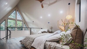 A bedroom with a sloped ceiling, large windows, a bed with a headboard, a hanging light fixture, and a staircase leading to an outdoor area. - Secluded A Frame Retreat,hot tub,sauna (Wright City)