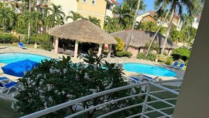 Uma área de piscina com espreguiçadeiras e guarda-sóis de palha, cercada por vegetação e edifícios.