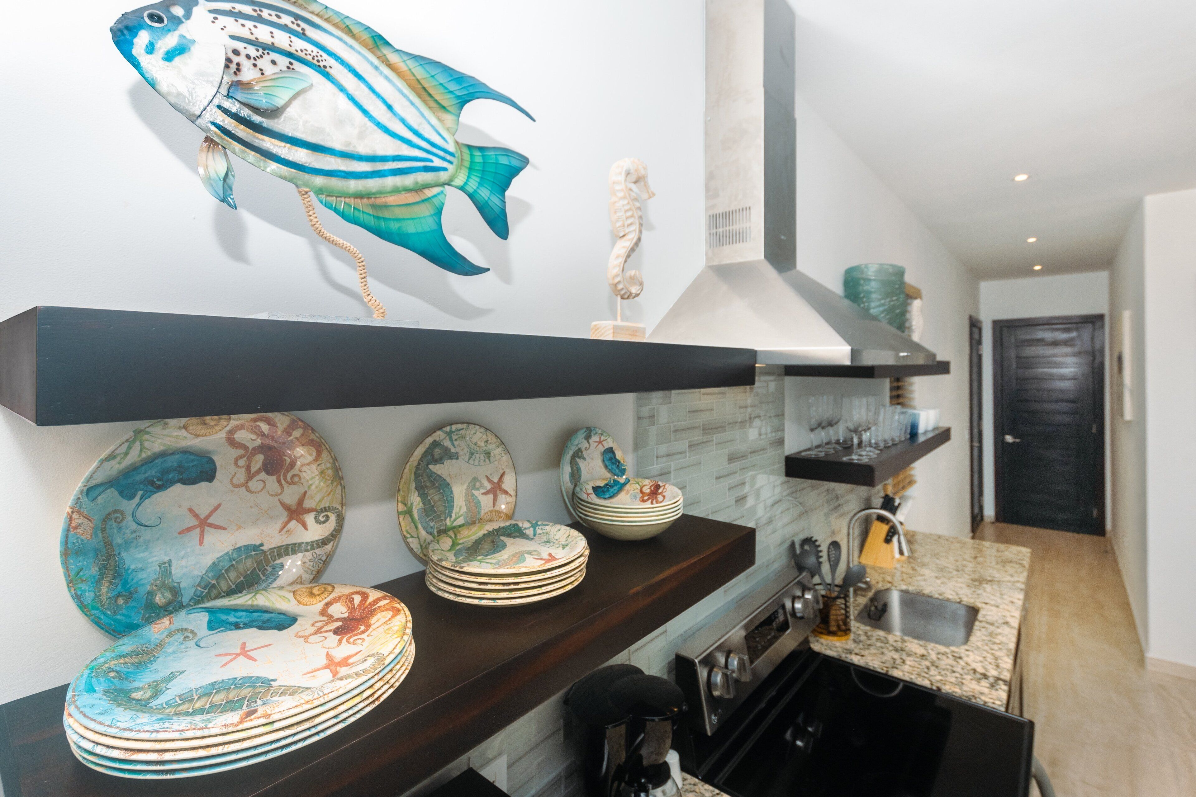Une cuisine avec un plan de travail en bois foncé, une décoration représentant un poisson accrochée au mur, et une variété d’assiettes et de bols disposés sur les étagères.