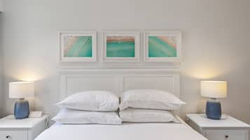 Une chambre à coucher bien rangée, avec un lit blanc, deux tables de chevet dotées de lampes et trois œuvres d’art encadrées au-dessus du lit.