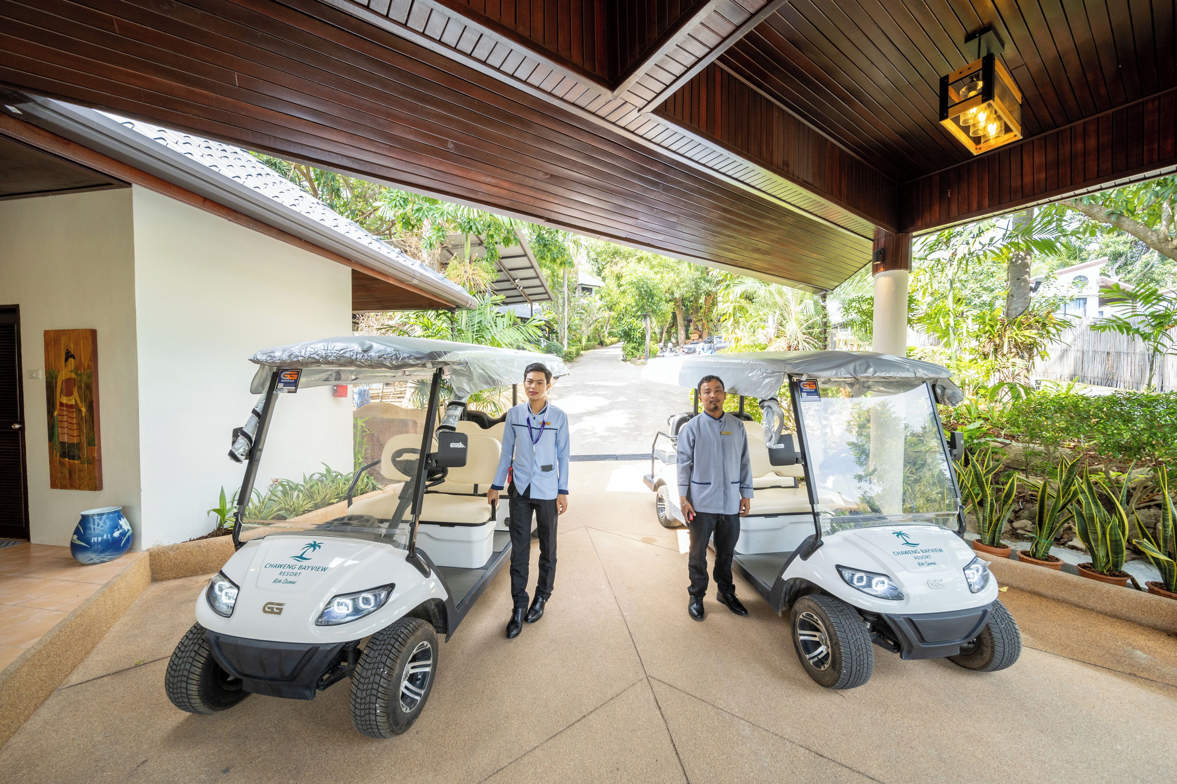 Two golf carts parked under a covered area with a wooden ceiling and potted plants around.