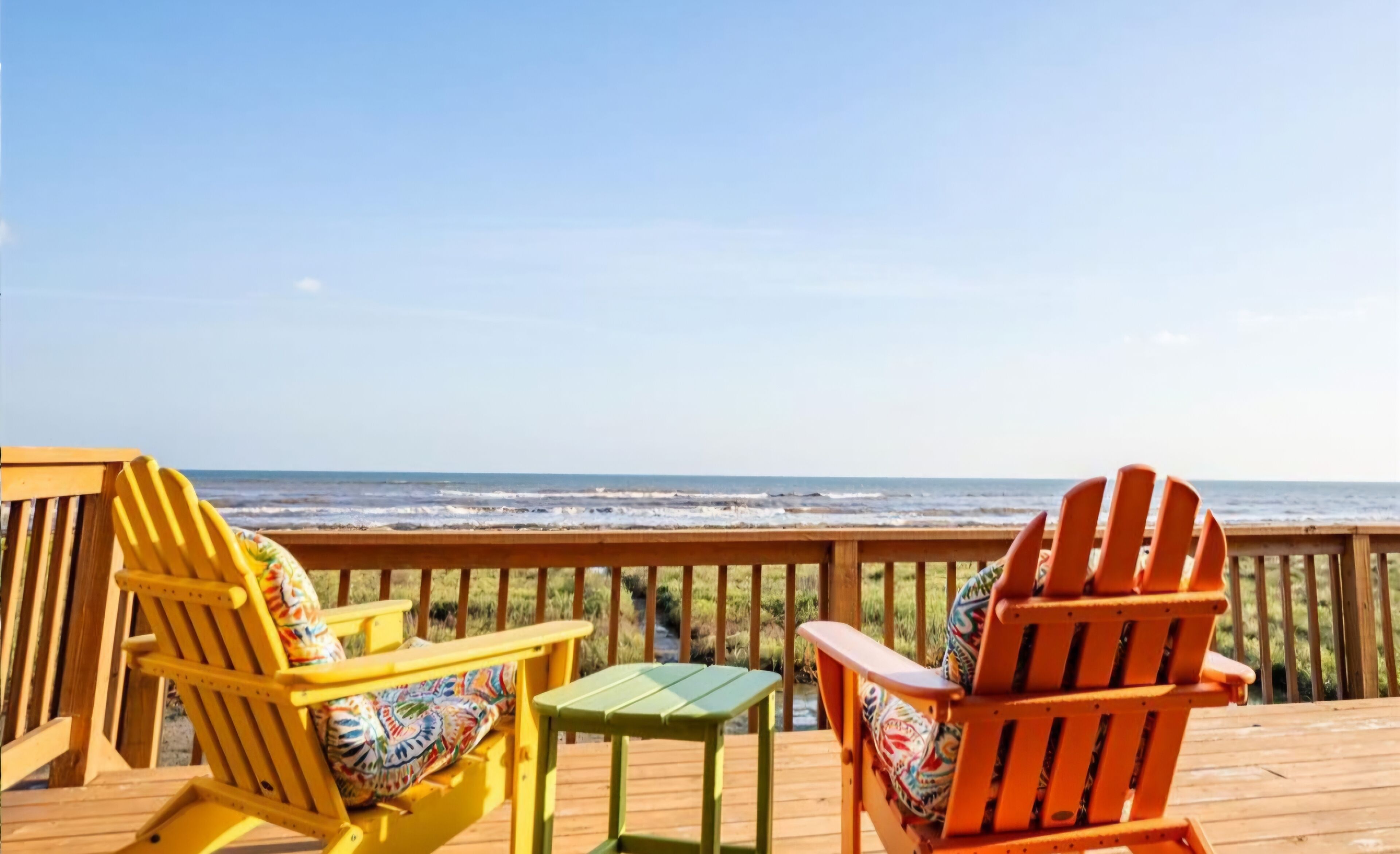 A wooden deck with two Adirondack chairs and a small table, overlooking a beach with waves.