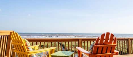 A wooden deck with two Adirondack chairs and a small table, overlooking a beach with waves.