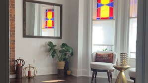 A spacious living room with hardwood floors, a large mirror, a potted plant, a comfortable armchair, and a window with a stained glass design. - Bright & Spacious Home close to NYC and World Cup (Bayonne)