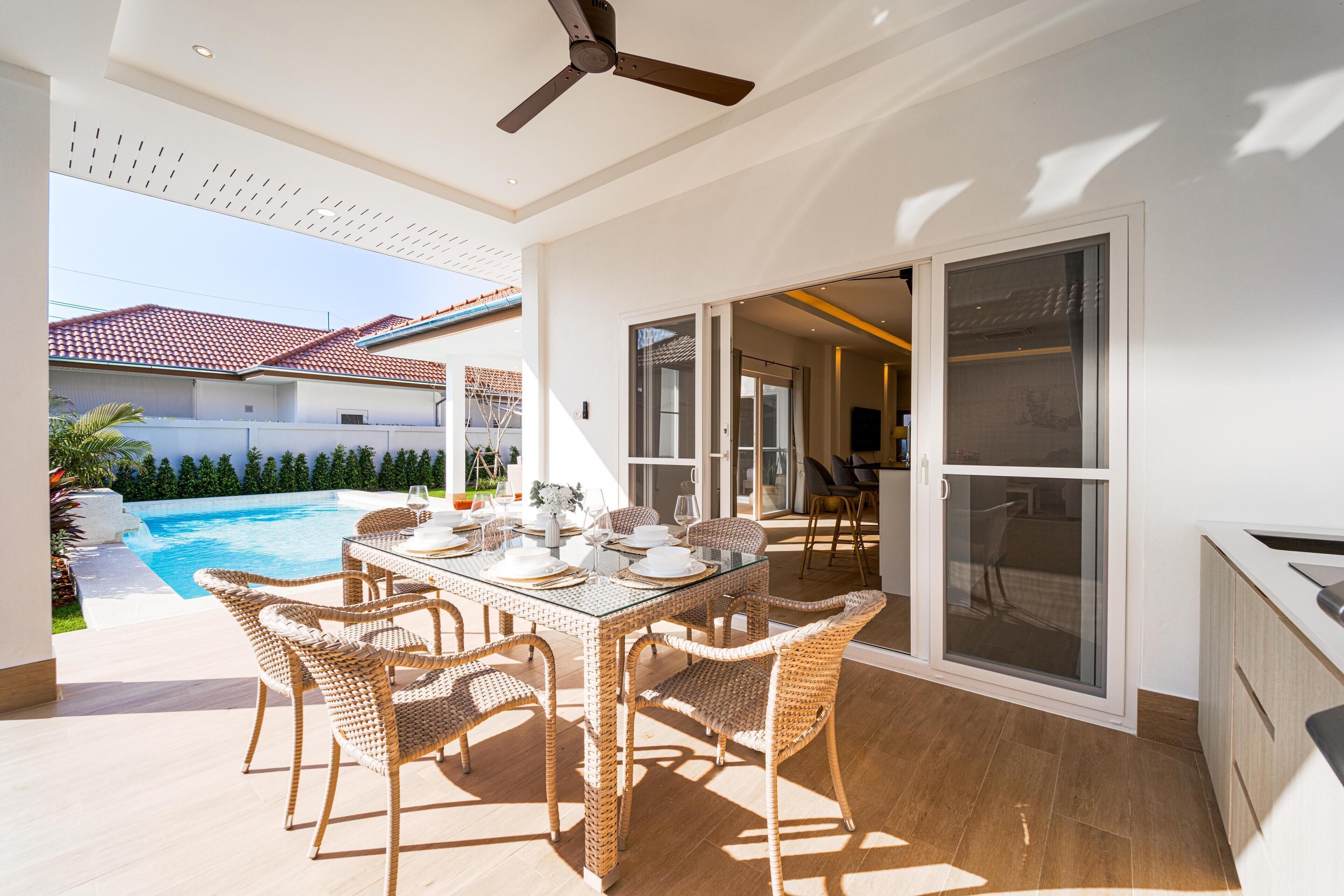 A modern kitchen with a large island, a dining area with wicker chairs, and a pool in the background.