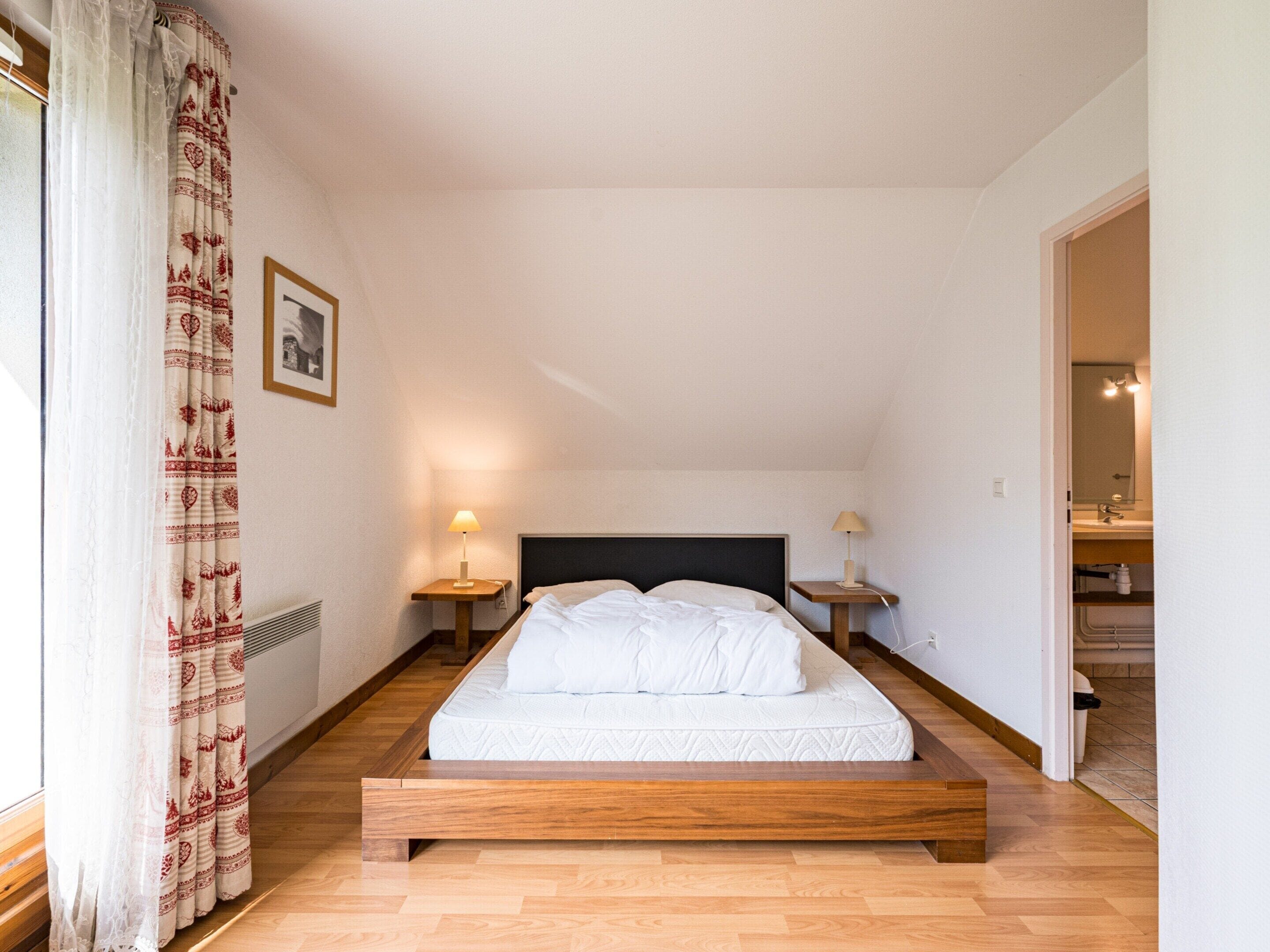 A bedroom with a wooden bed, two bedside tables with lamps, a radiator, and a window with patterned curtains.