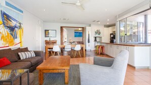 A modern living room with a wooden coffee table, a grey armchair, and a brown sofa. There is a dining area with a wooden table and chairs, and a kitchen with white cabinets and a large window. - Sunset Views walking distance to Fremantle (Fremantle)