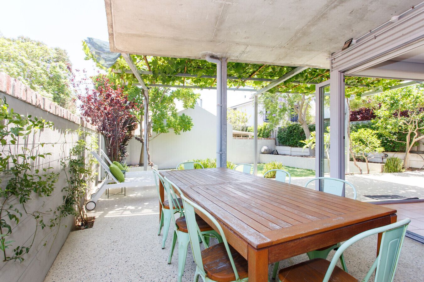 Een terras buiten met een houten tafel en groene stoelen, omringd door planten en een witte muur.