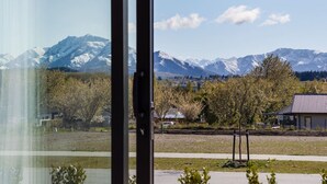 Blick durch ein Fenster auf eine Bergkette im Hintergrund, eine grasbewachsene Fläche mit Bäumen und ein Gebäude mit geneigtem Dach.