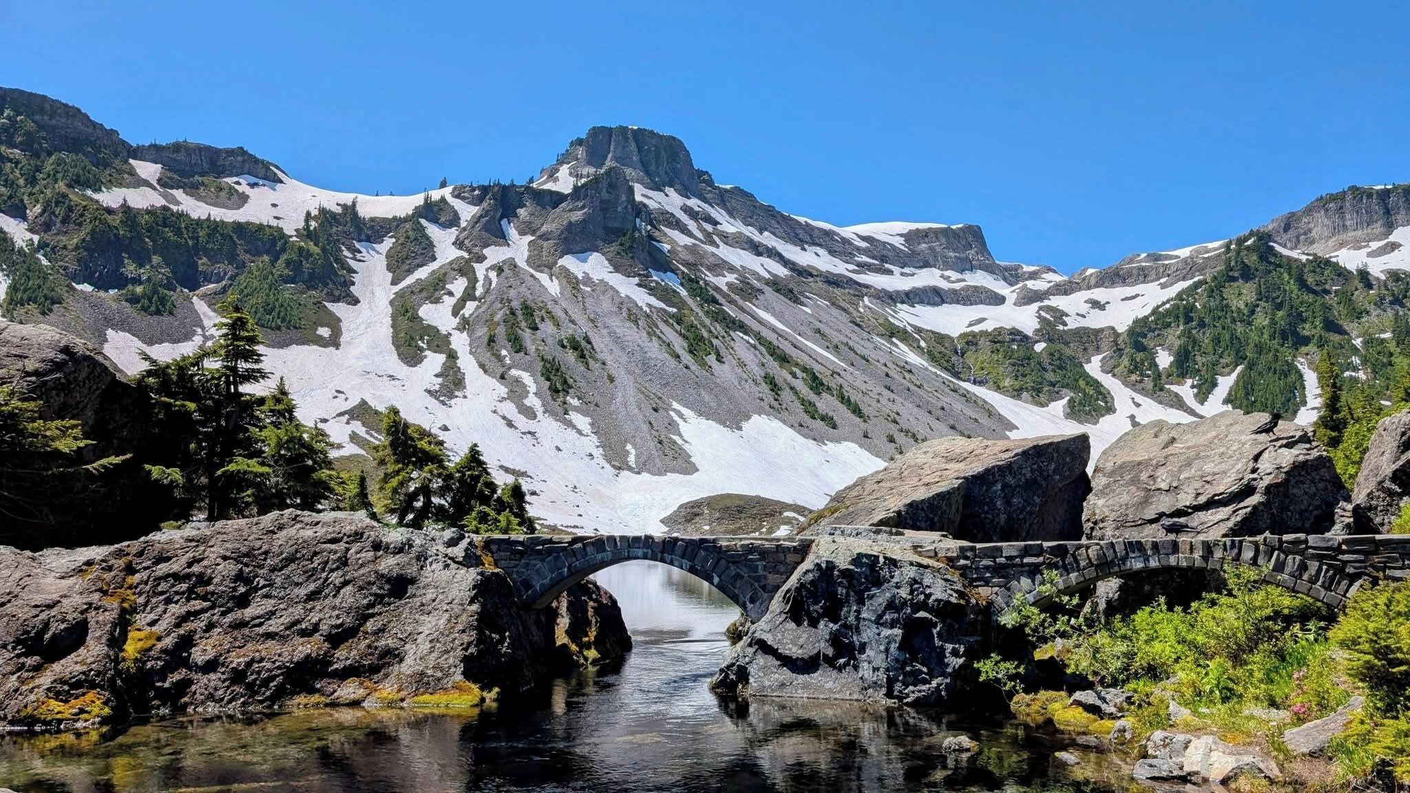 A mountain landscape with a stone bridge over a calm river, surrounded by snow-capped peaks and dense forest.
